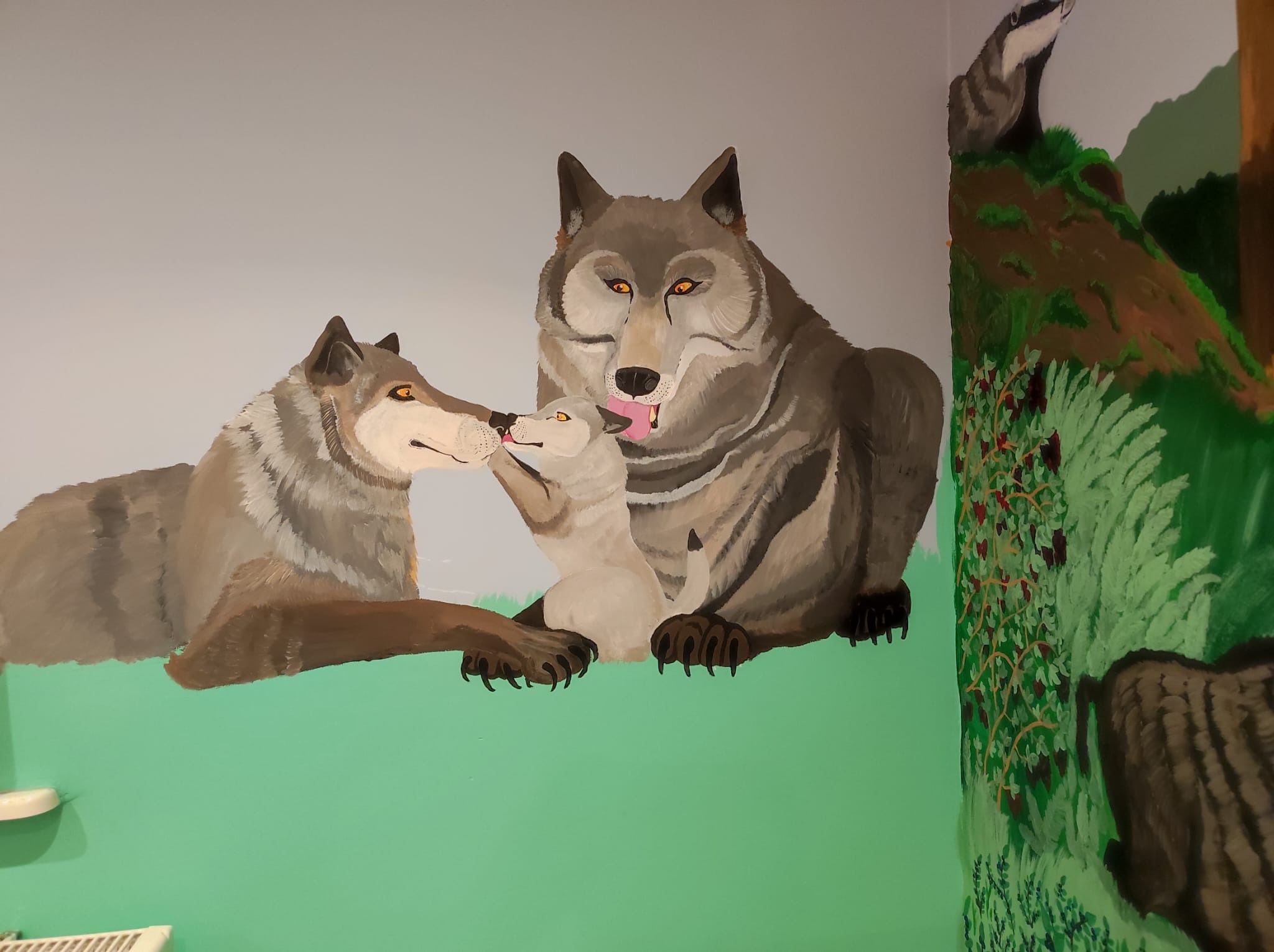 Naścienne malowidło przedstawia rodzinę wilków: dorosłego wilka, wilczycę i szczeniaka, otoczonych zielonym krajobrazem z elementami drzew i krzewów.