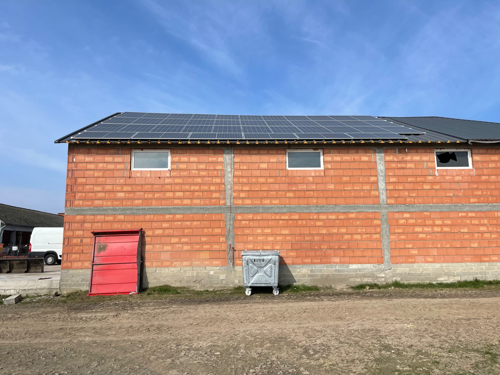 Budynek w trakcie budowy z zamontowanymi panelami fotowoltaicznymi na dachu, widoczne okna, czerwone drzwi i kontener na śmieci.