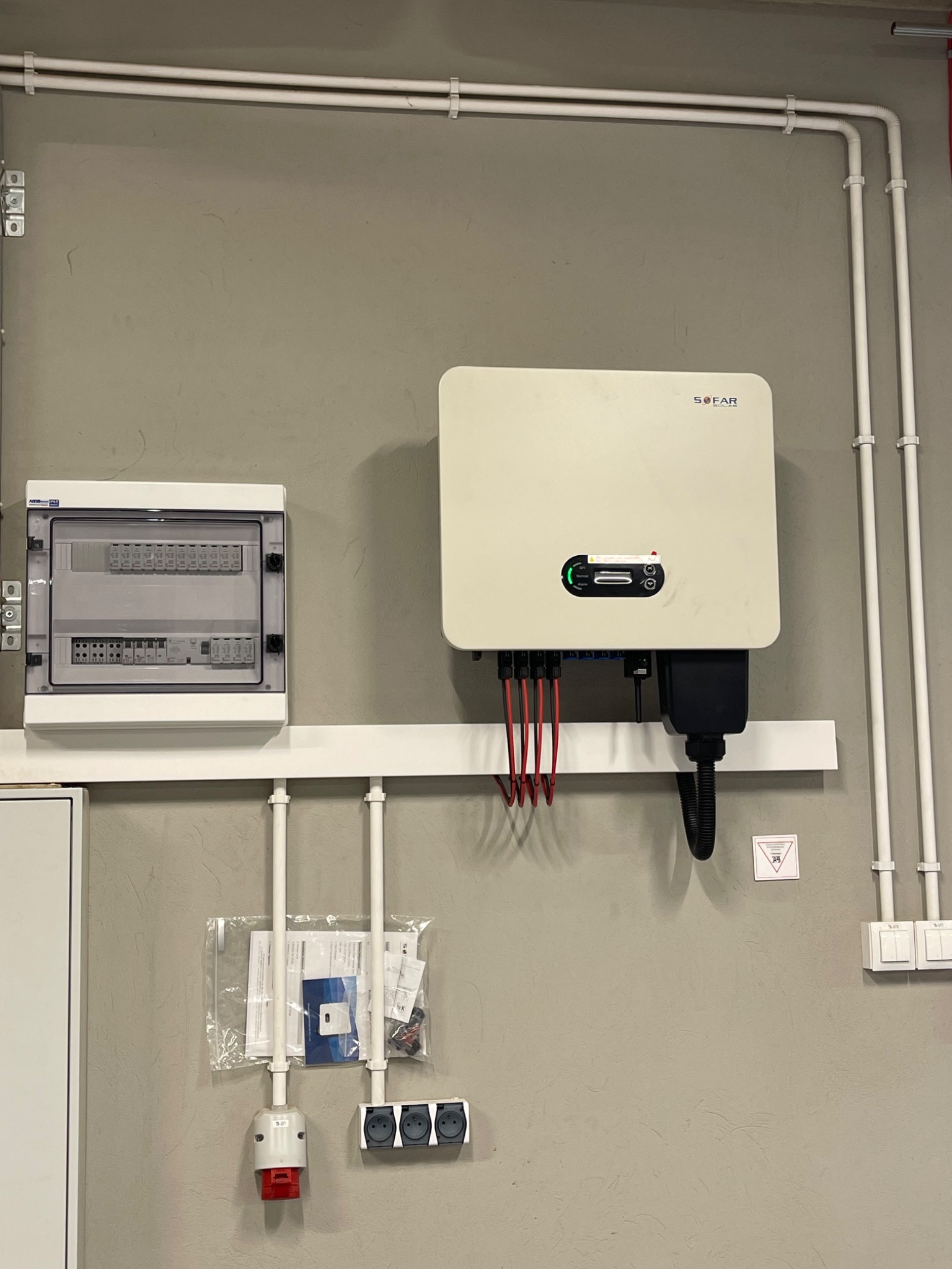 Biały inwerter SOFAR zamontowany na ścianie, z widocznymi przewodami, obok skrzynka elektryczna i gniazda, instalacja w szarej, surowej przestrzeni.