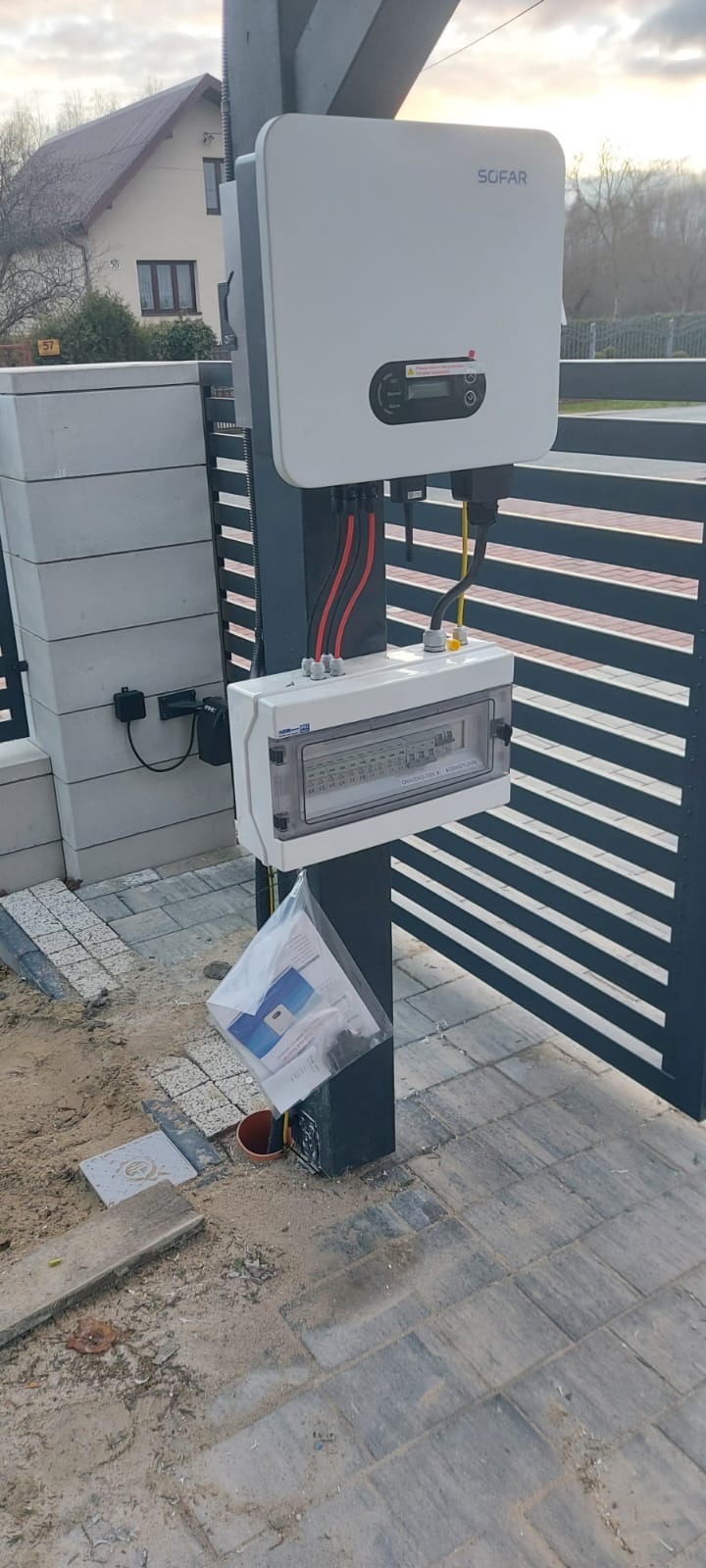 Biała skrzynka rozdzielcza z przewodami, zamontowana na słupku ogrodzenia, pod nią dokumentacja w plastikowej torbie, w tle dom jednorodzinny.