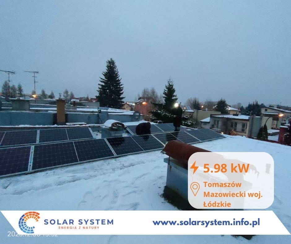 Instalacja paneli słonecznych na dachu pokrytym śniegiem w Tomaszowie Mazowieckim, pochmurny dzień, widoczny komin i drzewo iglaste w tle, logo firmy Solar System w lewym dolnym rogu.