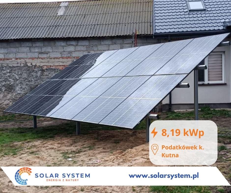 Instalacja fotowoltaiczna 8,19 kWp na gruncie, widoczna z boku na tle domu jednorodzinnego i budynku gospodarczego, częściowo pokryta rosą lub deszczem, z logo Solar System i adresem Podatcówek k.
