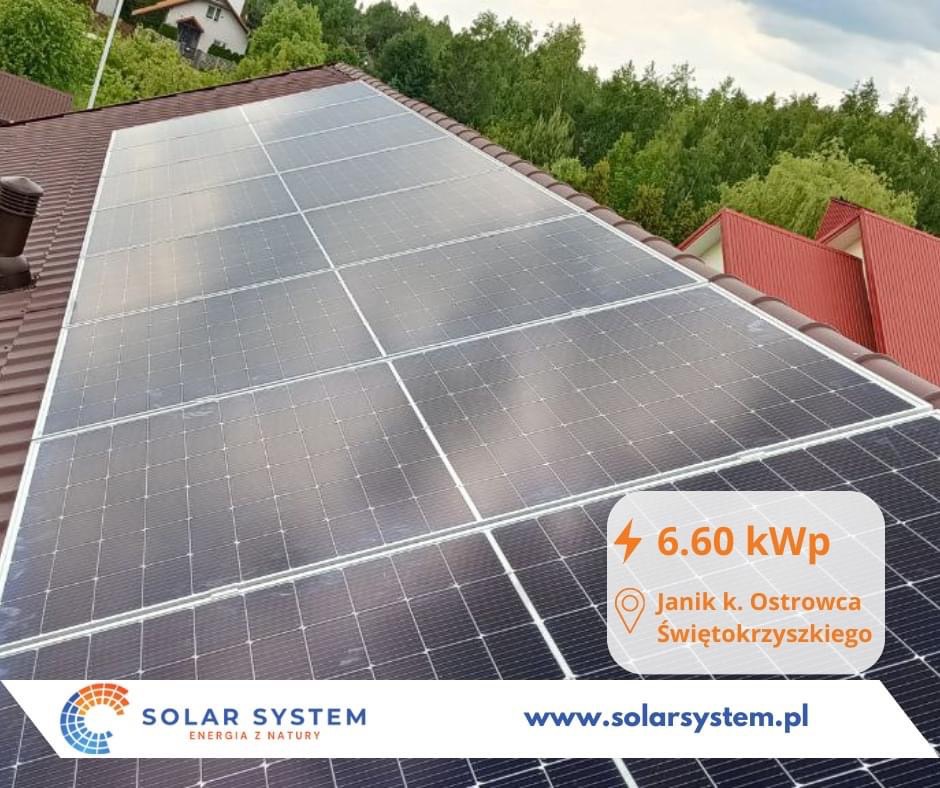 Panele słoneczne o mocy 6.60 kWp zamontowane na brązowym dachu domu jednorodzinnego, widok z góry na tle zieleni drzew, z widocznym logo firmy Solar System i adresem Janik k. Ostrowca...