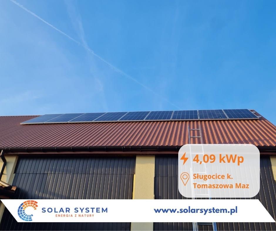 Panele słoneczne na brązowym dachu budynku z ciemnymi bramami garażowymi, widoczne logo firmy Solar System i informacja o mocy 4,09 kWp oraz lokalizacji Sługocice k. Tomaszowa Maz.