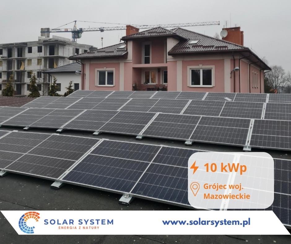 Panele słoneczne o mocy 10 kWp na dachu dwupiętrowego różowego domu w Grójcu, woj. Mazowieckie, z budową w tle i logo firmy Solar System w rogu.