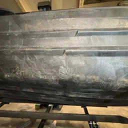 Spód czarnej łodzi lub kajaka z widocznymi śladami użytkowania i napraw, umieszczony na metalowym stojaku.