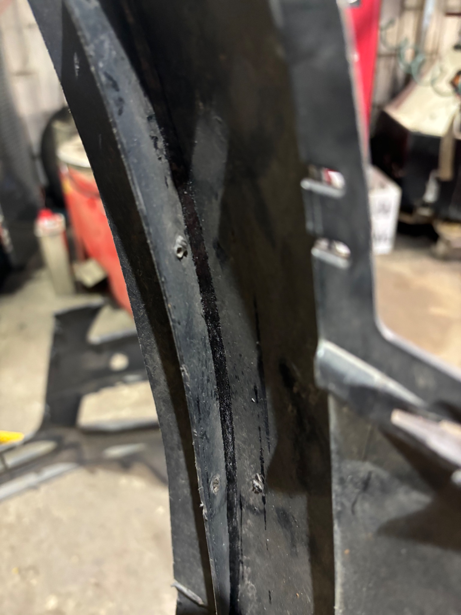 Fragment czarnego, plastikowego zderzaka samochodowego z widocznymi otworami i śladami naprawy, w tle warsztat.