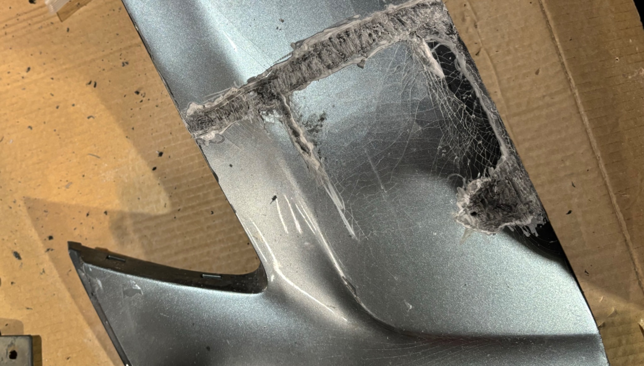 Uszkodzony, srebrny element karoserii samochodu po spawaniu, widoczne ślady spawu i pęknięcia wypełnione spoiwem.