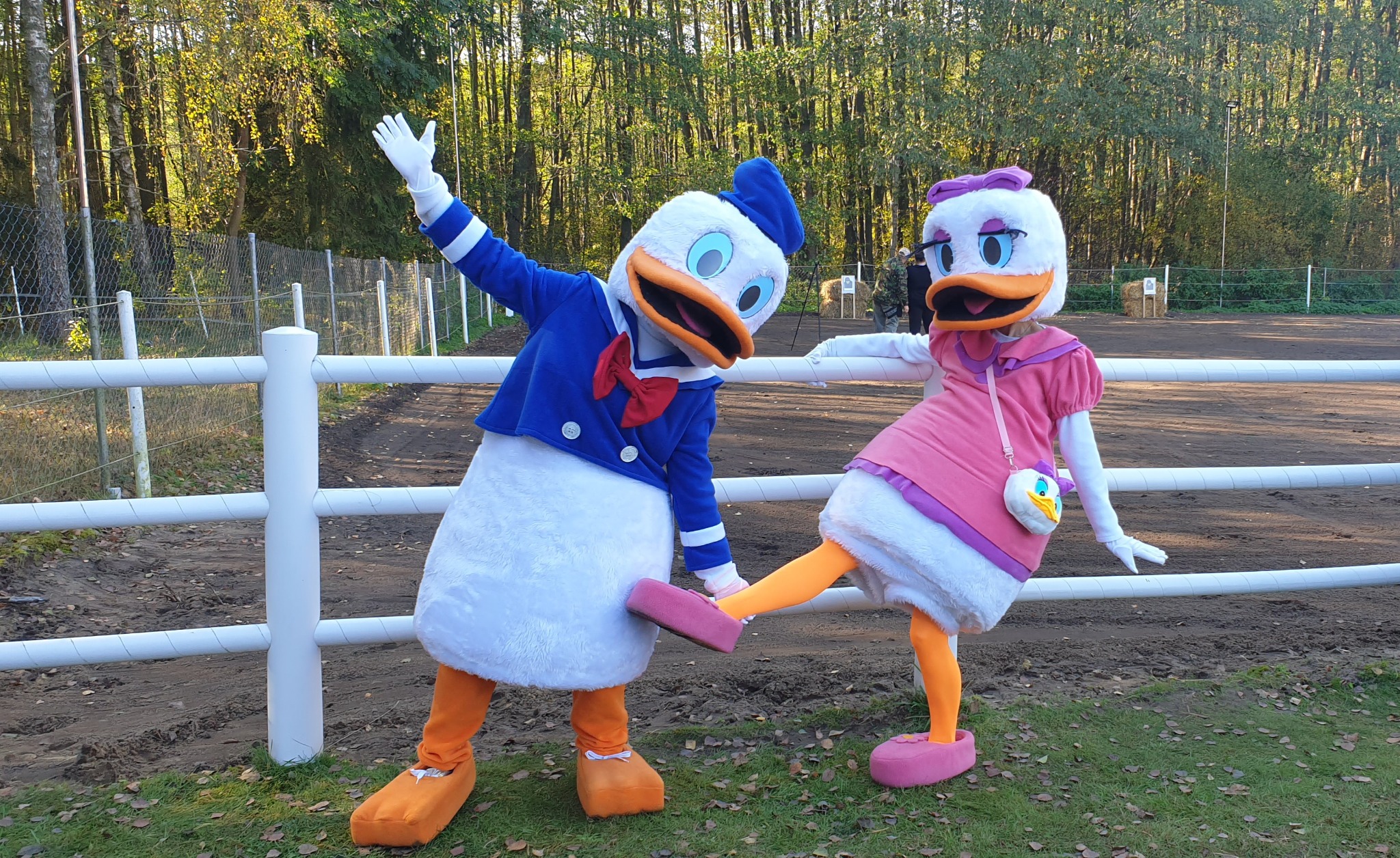 Dwie osoby w kostiumach Kaczora Donalda i Daisy Duck pozują przed białym płotem na tle drzew.
