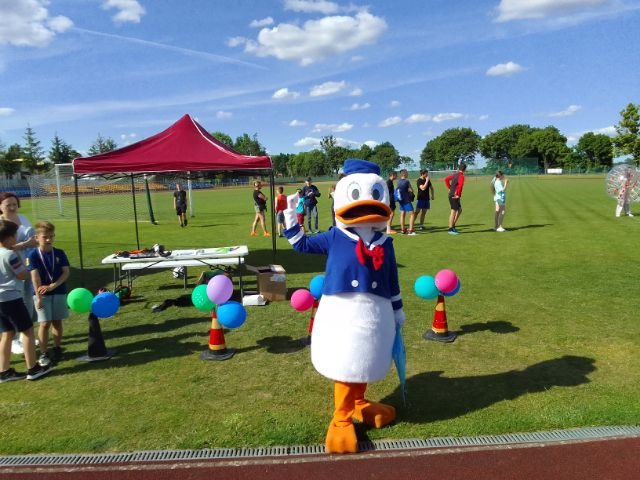 Postać z bajki Kaczor Donald w kostiumie na zielonym boisku sportowym w otoczeniu dzieci i rekwizytów animacyjnych.