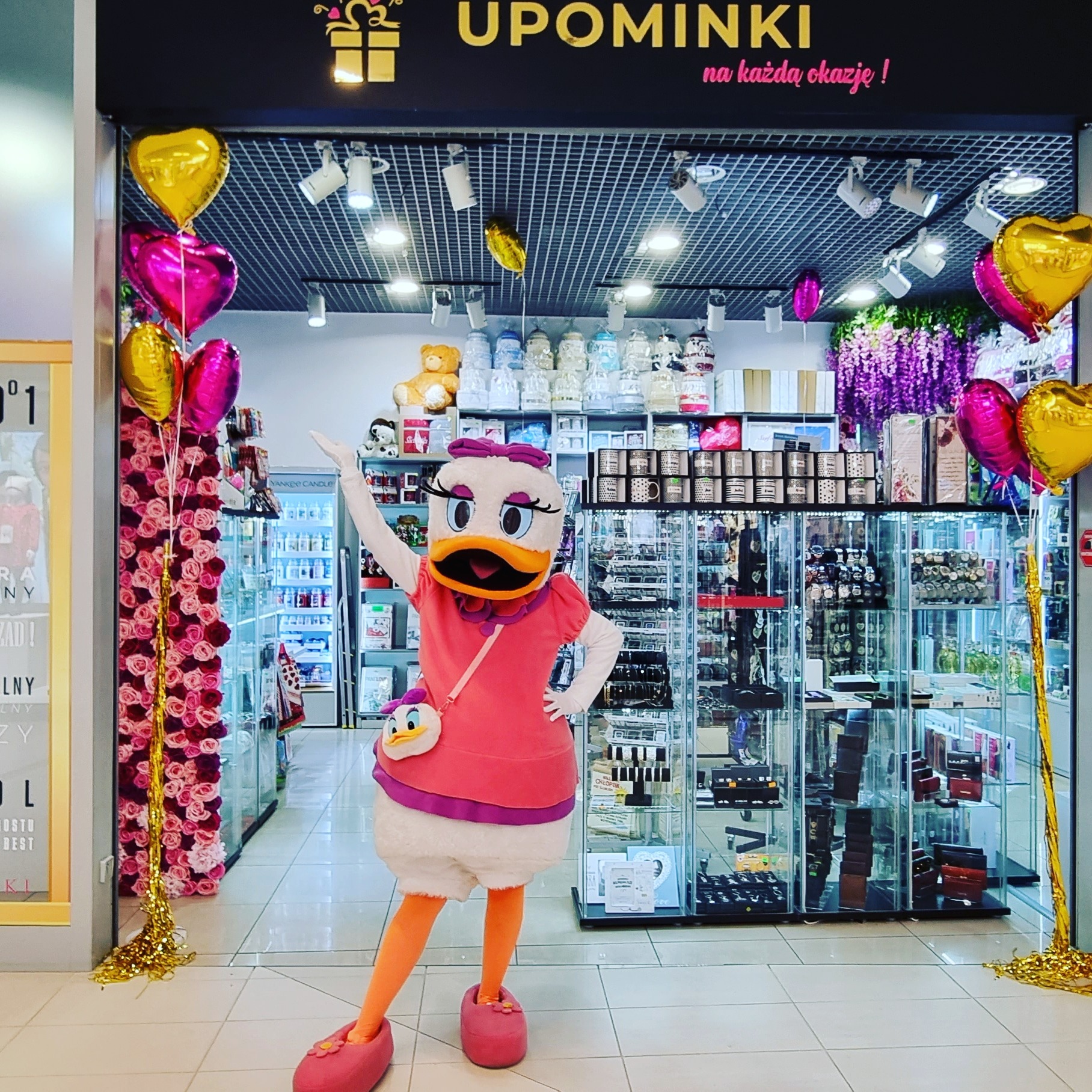 Osoba w kostiumie Daisy Duck stoi w wejściu do sklepu z upominkami, udekorowanego balonami w kształcie serc i girlandami kwiatowymi.