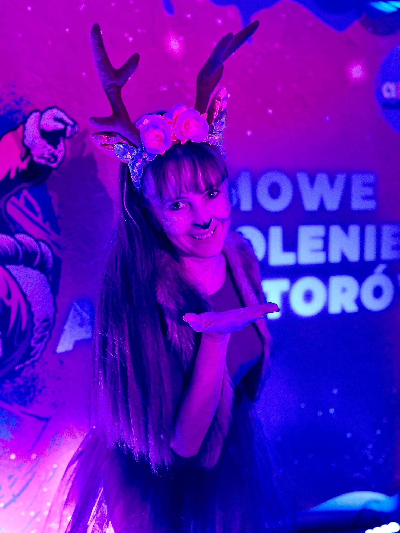 Kobieta w kostiumie renifera z kwiatowym wiankiem i pomalowanym nosem, dmuchająca w dłoń, na tle dekoracji eventowej z neonowym oświetleniem.