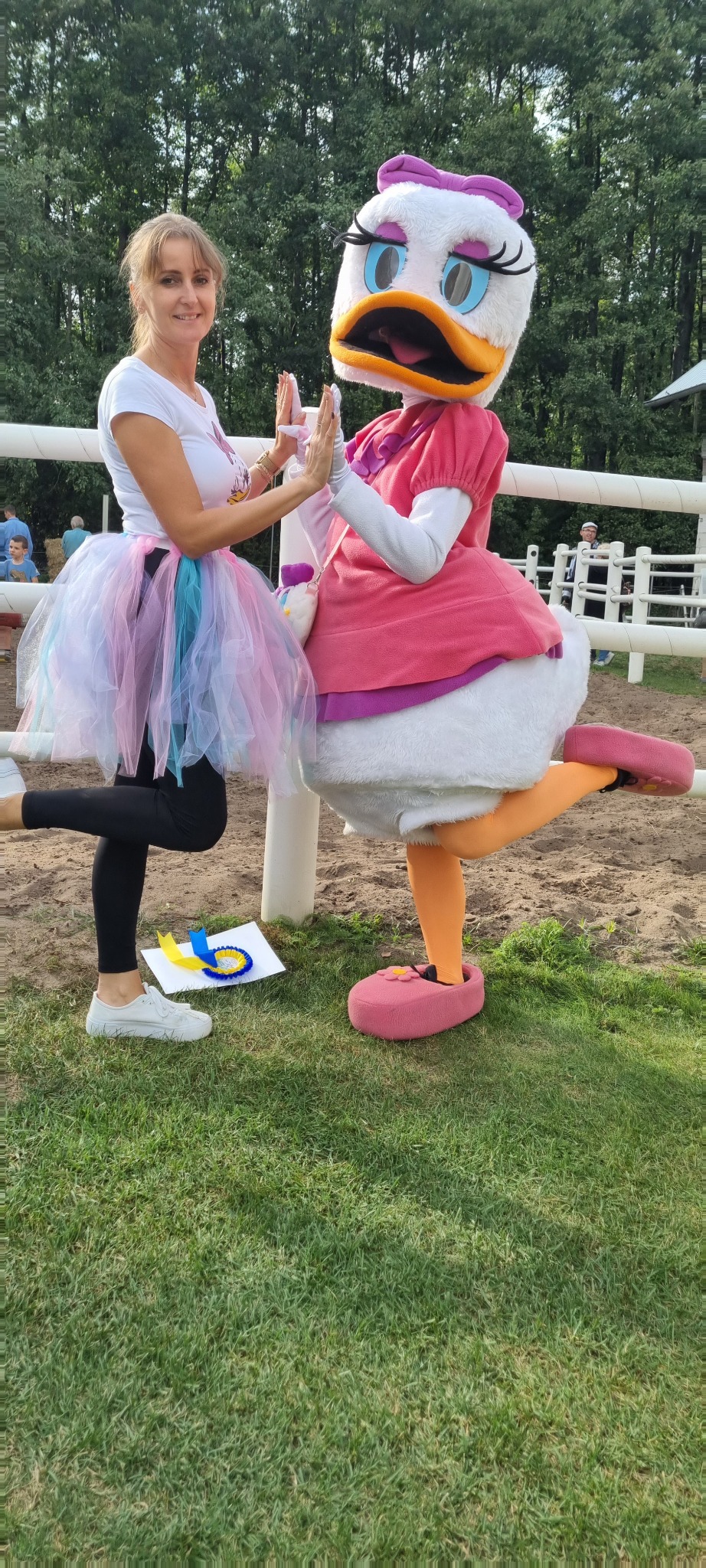 Kobieta w kolorowej tiulowej spódniczce przybija piątkę z osobą w kostiumie Kaczki Daisy na trawie, obok leży nagroda z niebiesko-żółtą wstążką.