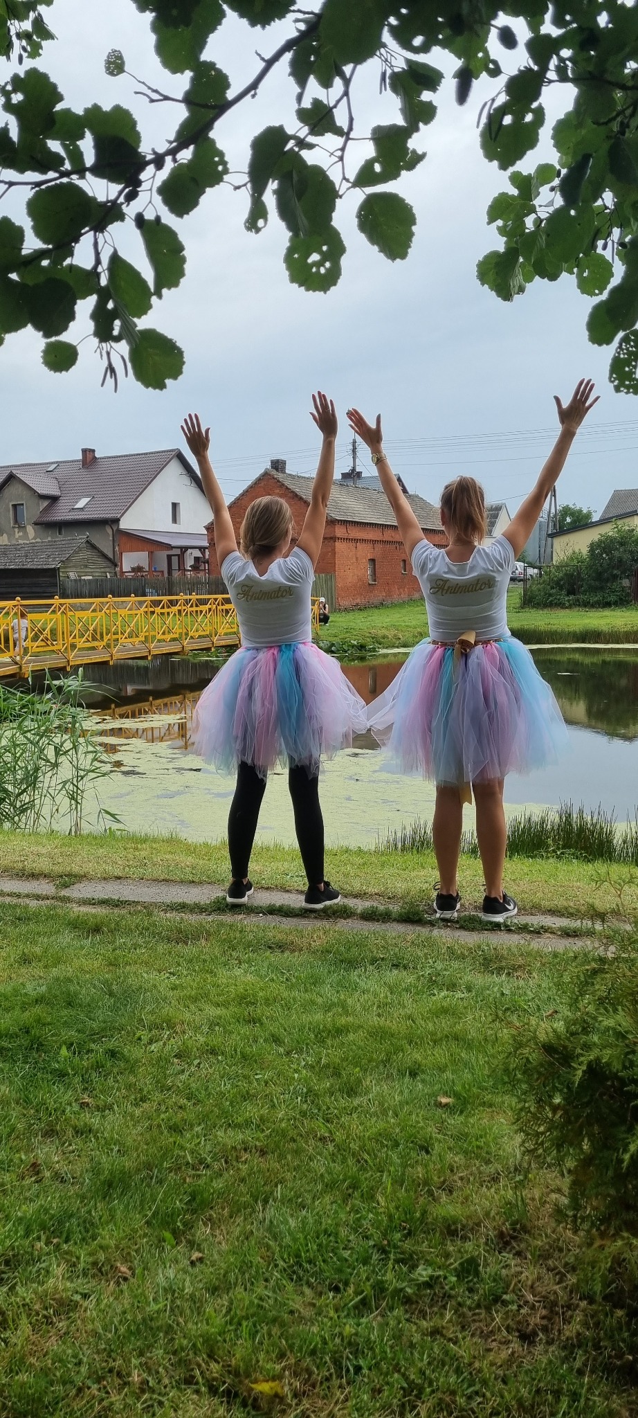 Dwie kobiety w tiulowych spódniczkach i koszulkach z napisem 'Animator' stoją tyłem z podniesionymi rękami na tle stawu i żółtego mostku.