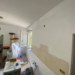 Wnętrze mieszkania w trakcie malowania: ściana z łatą i widocznymi śladami szpachlowania, wiadro z farbą, wałek i akcesoria malarskie na zabezpieczonej folią podłodze, drabina i drapak dla kota w tle.