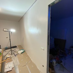 Boroda_Bud - Świeżo pomalowana na biało ściana w trakcie remontu, widoczny reflektor na statywie, zabezpieczona podłoga kartonem, oraz otwór drzwiowy prowadzący do pomieszczenia z niebieskim oświetleniem.