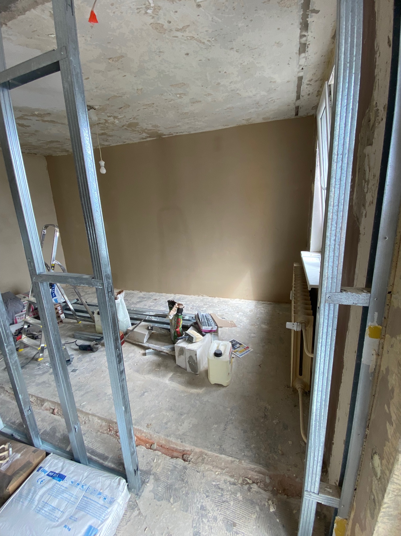 Pokój w trakcie prac wykończeniowych: widoczne metalowe profile konstrukcyjne, częściowo otynkowana ściana, drabina, narzędzia i materiały budowlane na podłodze. Sufit z widocznymi śladami...