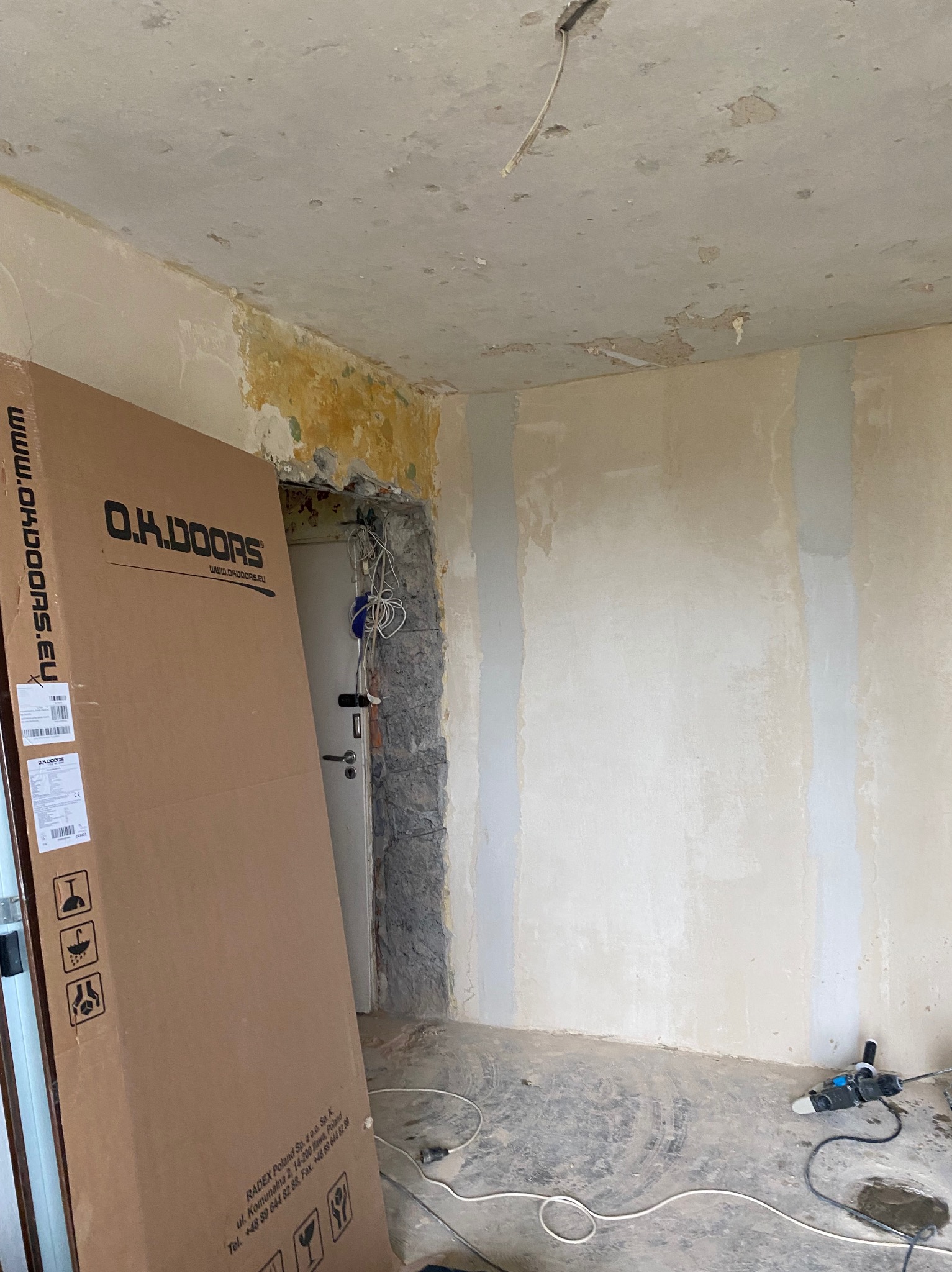 Wnętrze w trakcie remontu, widoczne przygotowane ściany do malowania, paczka drzwi O.K. Doors oparta o ścianę, oraz narzędzie do szlifowania na podłodze.