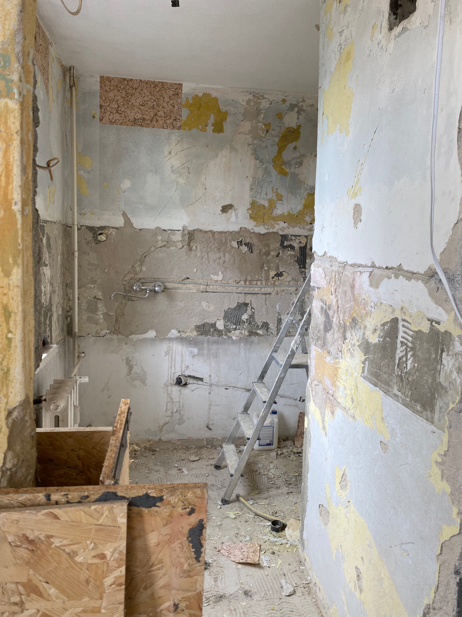 Pokój w trakcie remontu: zerwana tapeta, odsłonięte warstwy farby, drabina oparta o ścianę, widoczne instalacje wodne, fragment drewnianej konstrukcji.