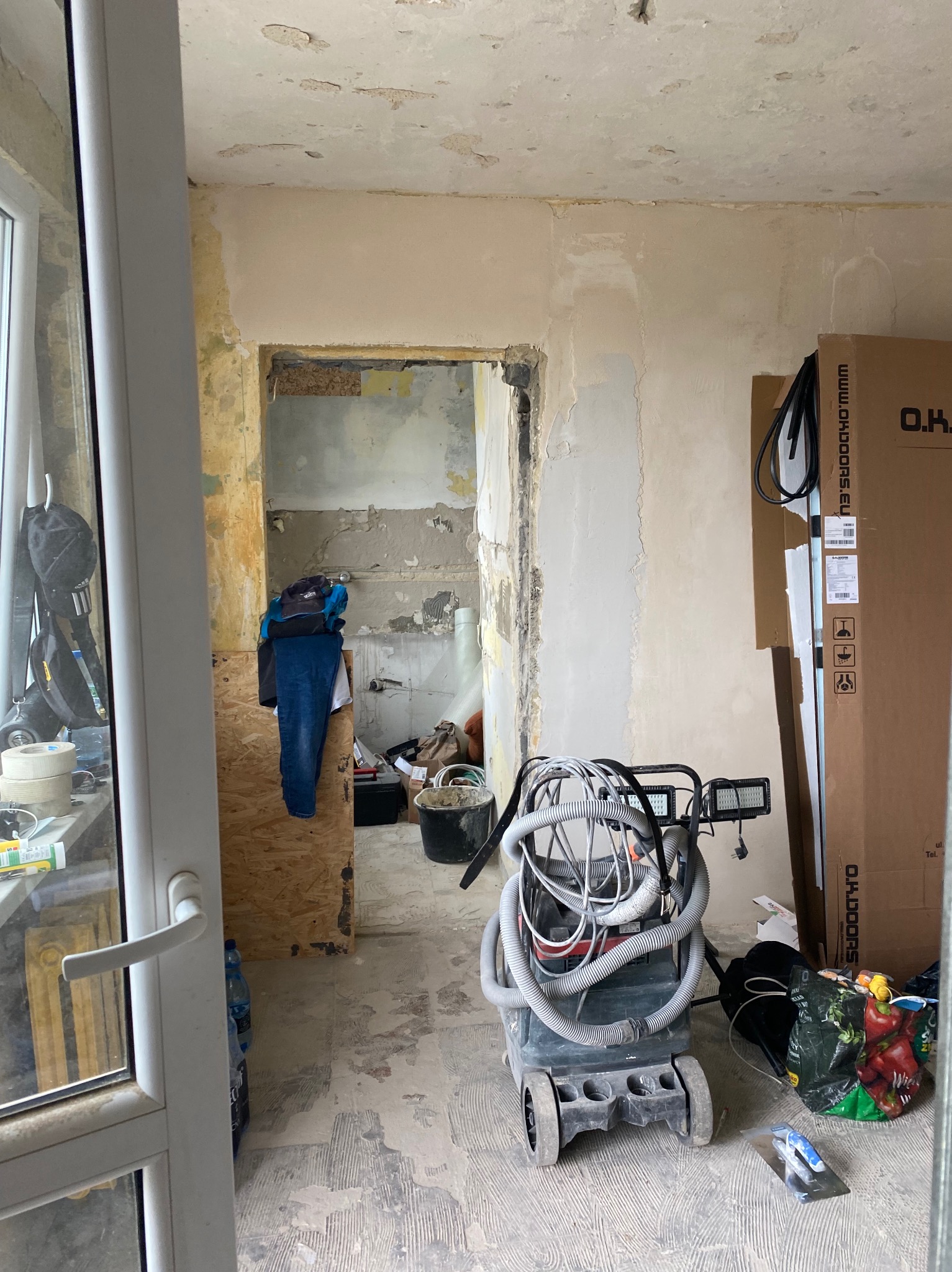 Wnętrze w trakcie remontu: szlifierka do gładzi z oświetleniem, odsłonięte ściany z resztkami farby i tapet, podłoga przygotowana do dalszych prac wykończeniowych, karton z drzwiami w tle.