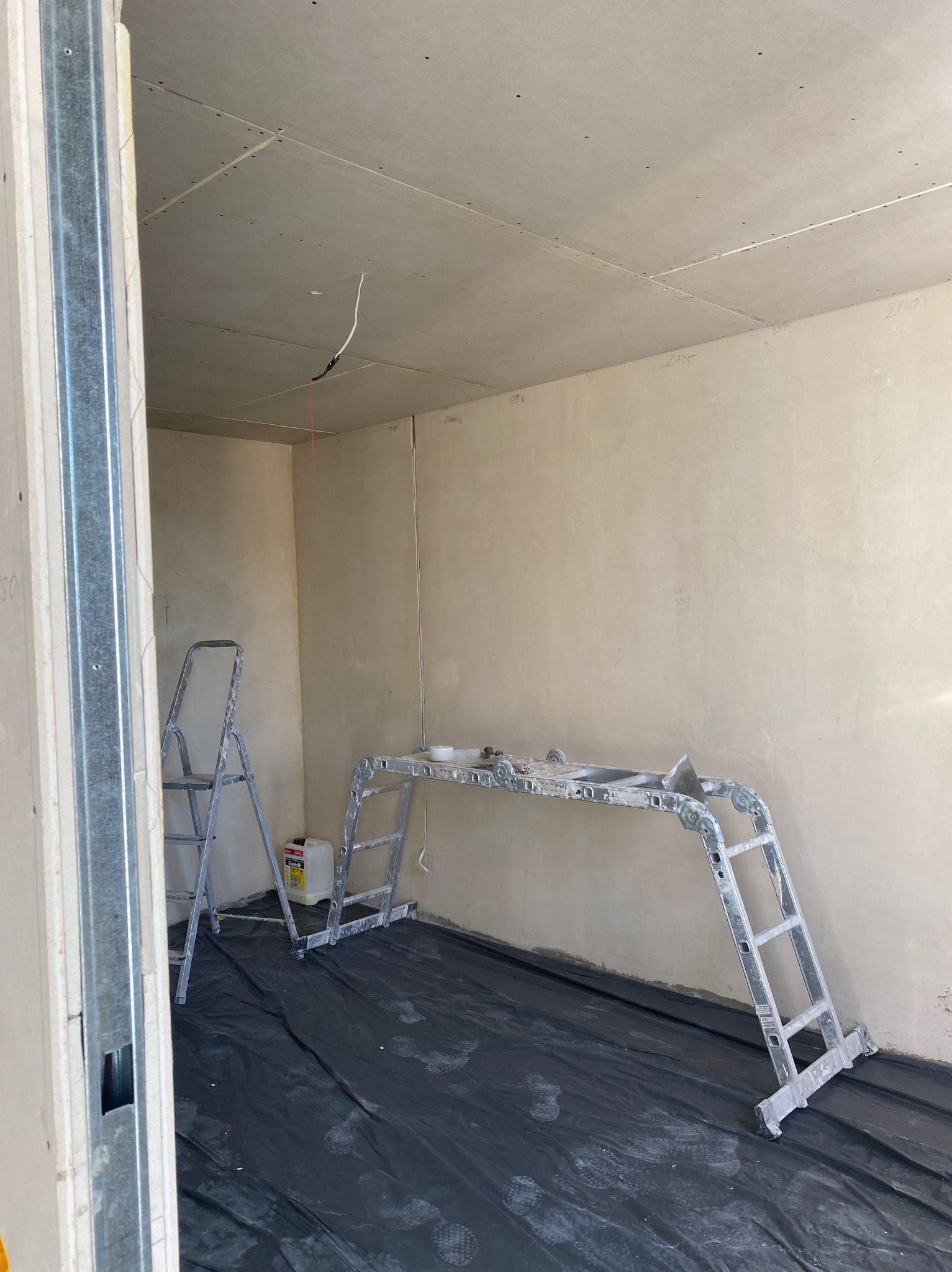 Pokój w trakcie szpachlowania ścian i sufitu, widoczny podest malarski, drabina, folia ochronna na podłodze i wystające kable elektryczne z sufitu.