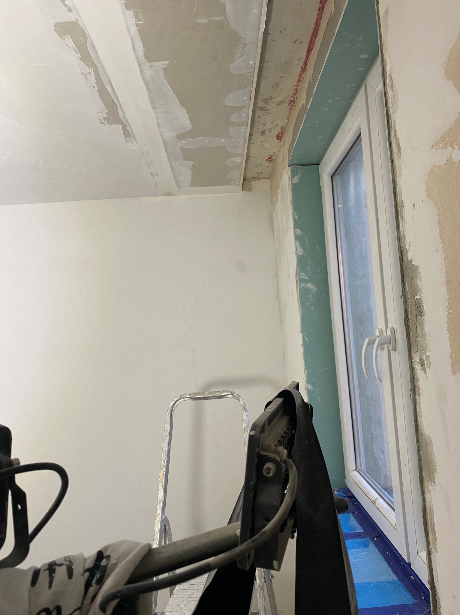 Pokój w trakcie prac wykończeniowych: szpachlowanie sufitu i ścian, zabezpieczone okno folią, widoczna drabina i lampa oświetleniowa.