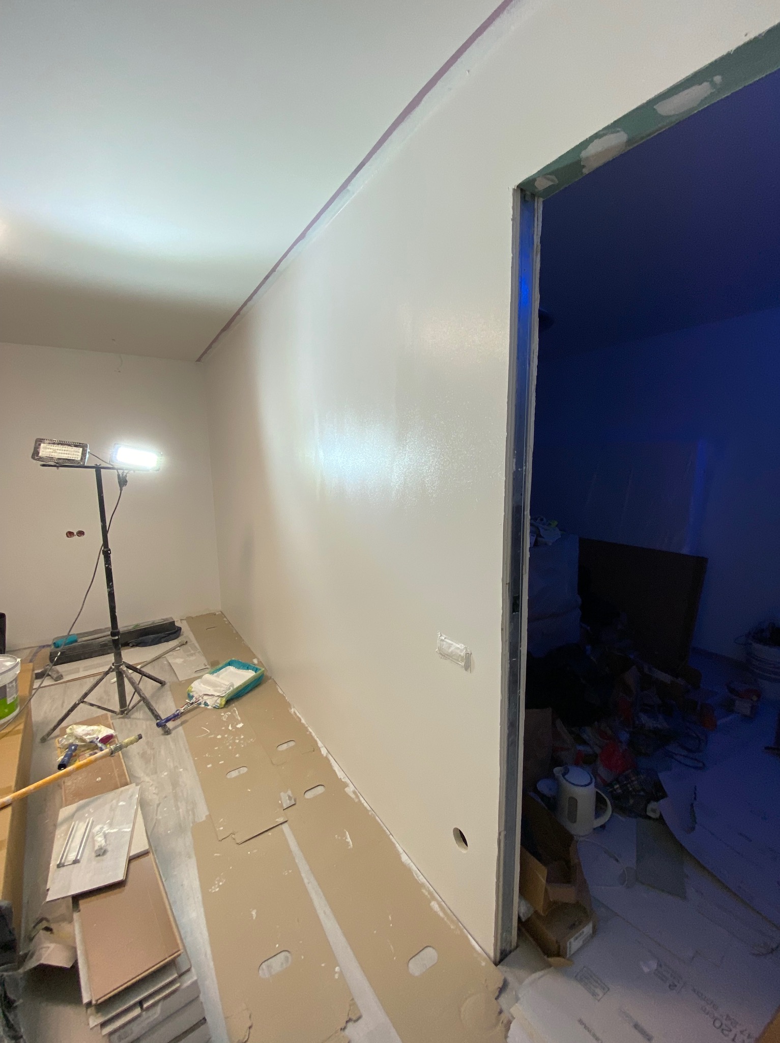 Świeżo pomalowana na biało ściana w trakcie remontu, widoczny reflektor na statywie, zabezpieczona podłoga kartonem, oraz otwór drzwiowy prowadzący do pomieszczenia z niebieskim oświetleniem.
