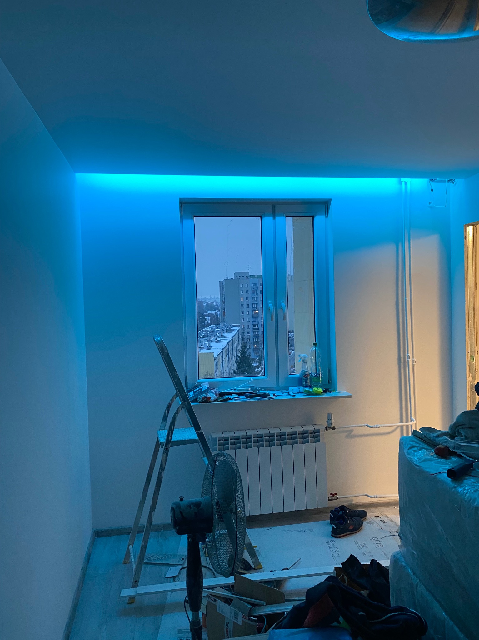 Pokój w trakcie prac wykończeniowych z zamontowanym oświetleniem LED podsufitowym, drabina oparta o ścianę, widoczny grzejnik, wentylator i okno z widokiem na blokowisko.