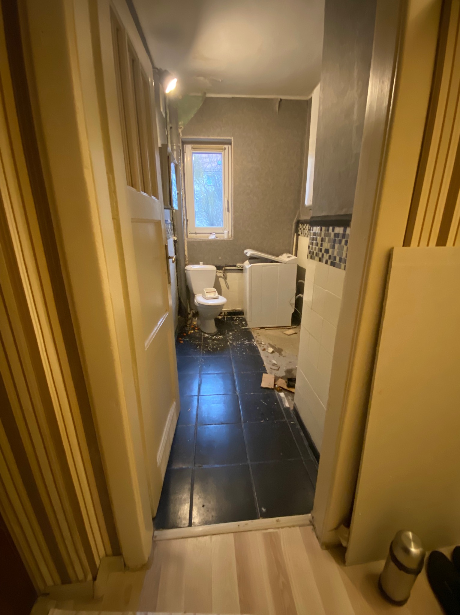 Łazienka w trakcie demontażu, widoczne odchylone drzwi, częściowo zdemontowana toaleta, płytki i pralka, pozostałości gruzu na podłodze. Widok z perspektywy wejścia.