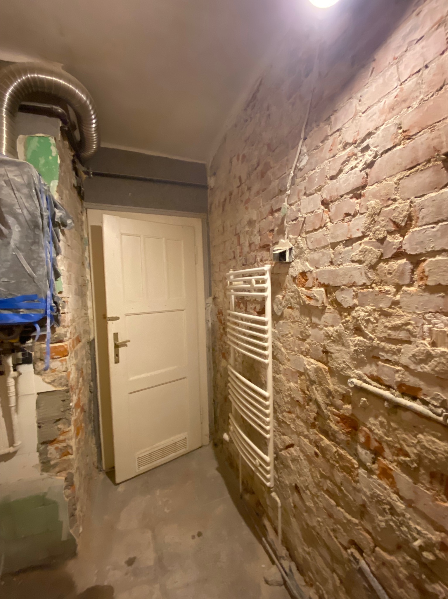 Wnętrze w trakcie remontu: odsłonięta ceglana ściana, biały grzejnik drabinkowy, białe drzwi i widoczne instalacje gazowe.