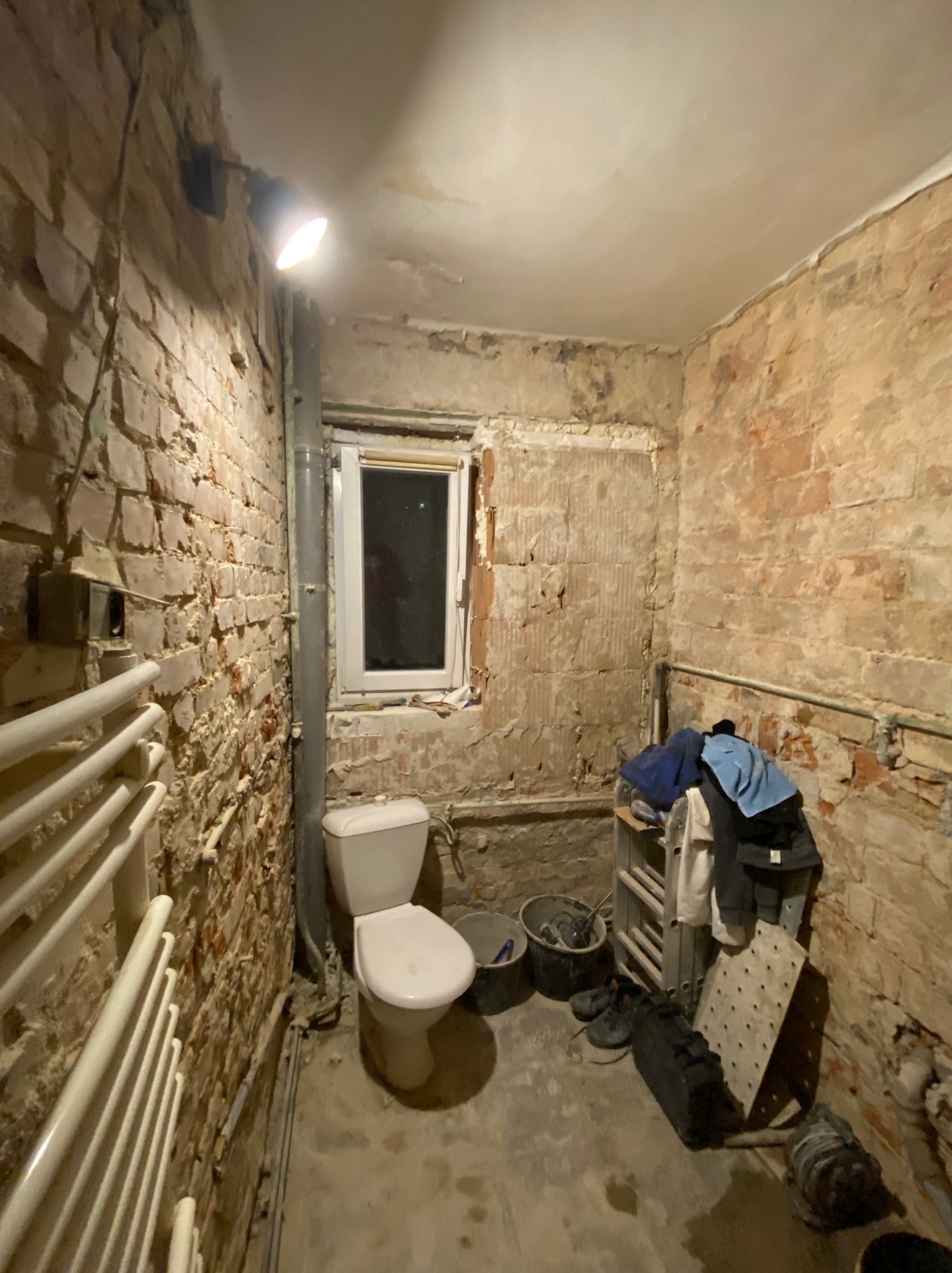 Łazienka w trakcie remontu: odsłonięta cegła, surowy beton, wisząca lampa halogenowa, toaleta, grzejnik drabinkowy, wiadro z narzędziami i prowizoryczny regał z ubraniami.