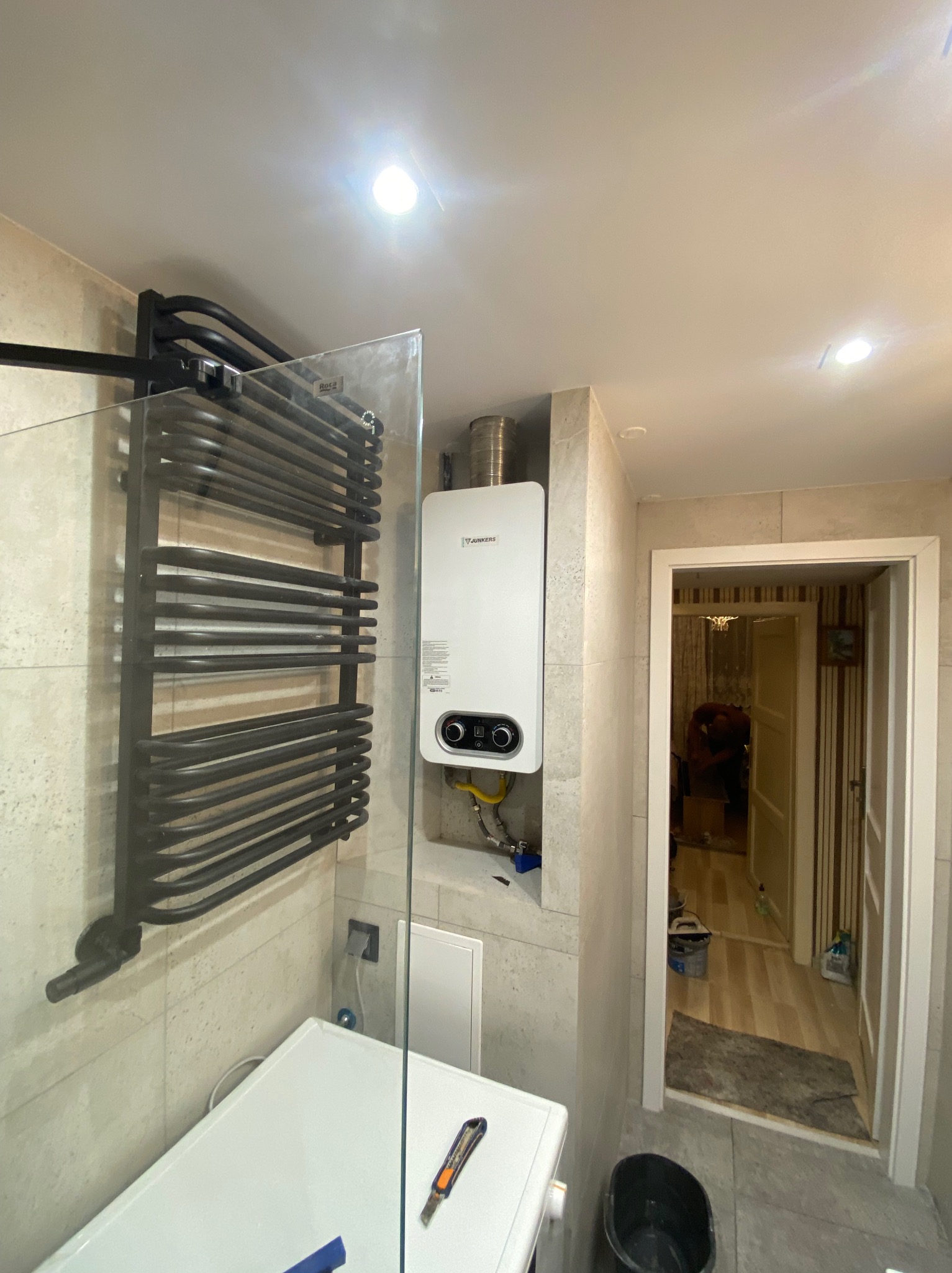 Nowoczesna łazienka w trakcie remontu: biały piec gazowy Junkers z podłączeniem, czarny grzejnik drabinkowy na ścianie z beżowych płytek, widoczna pralka i narzędzie do cięcia, otwarte drzwi...