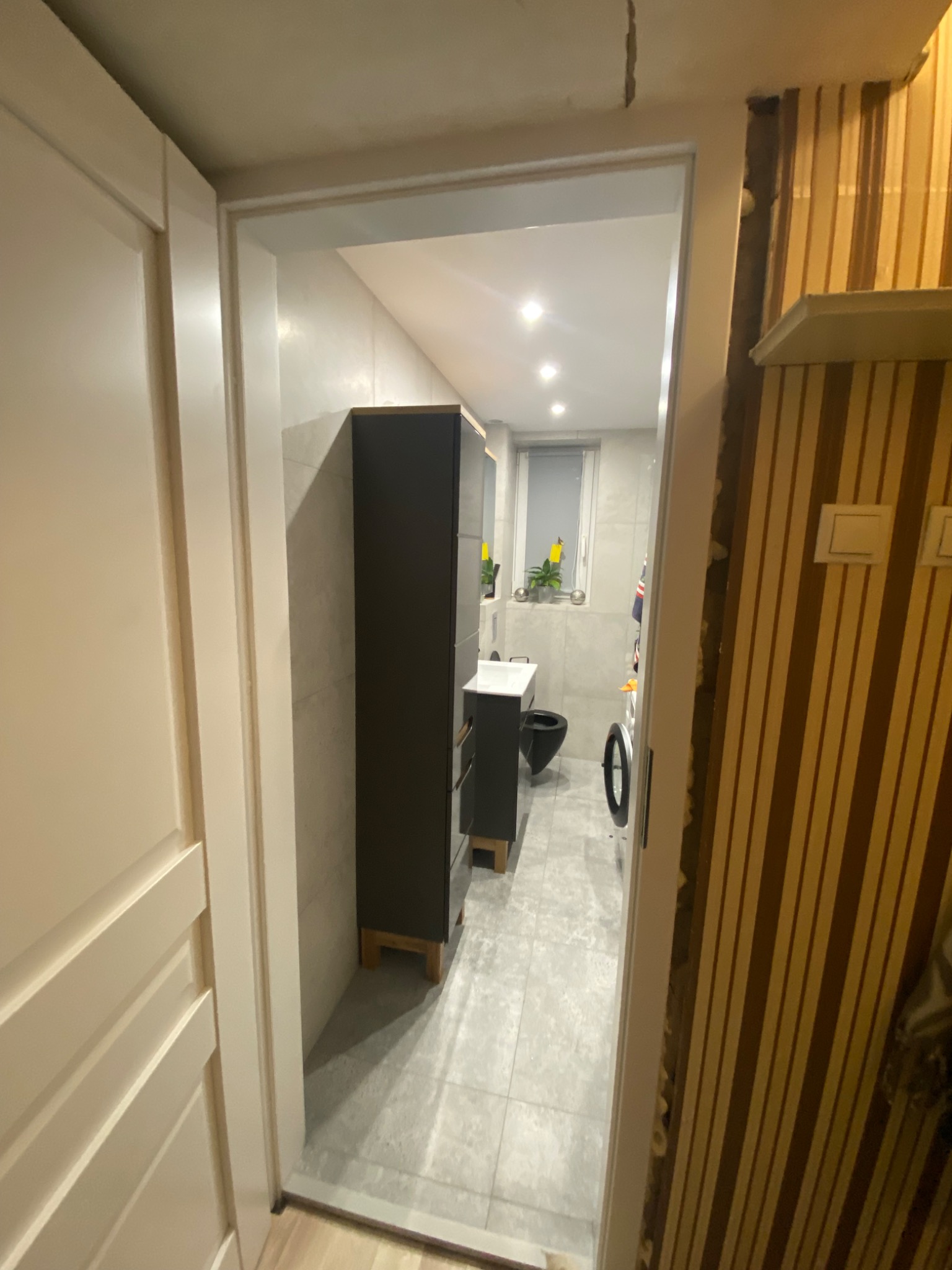 Widok przez futrynę na wyremontowaną łazienkę z szarą podłogą i grafitowymi meblami, gdzie po prawej stronie widać fragment ściany z tapetą w brązowe pasy i niedokończonymi wykończeniami.