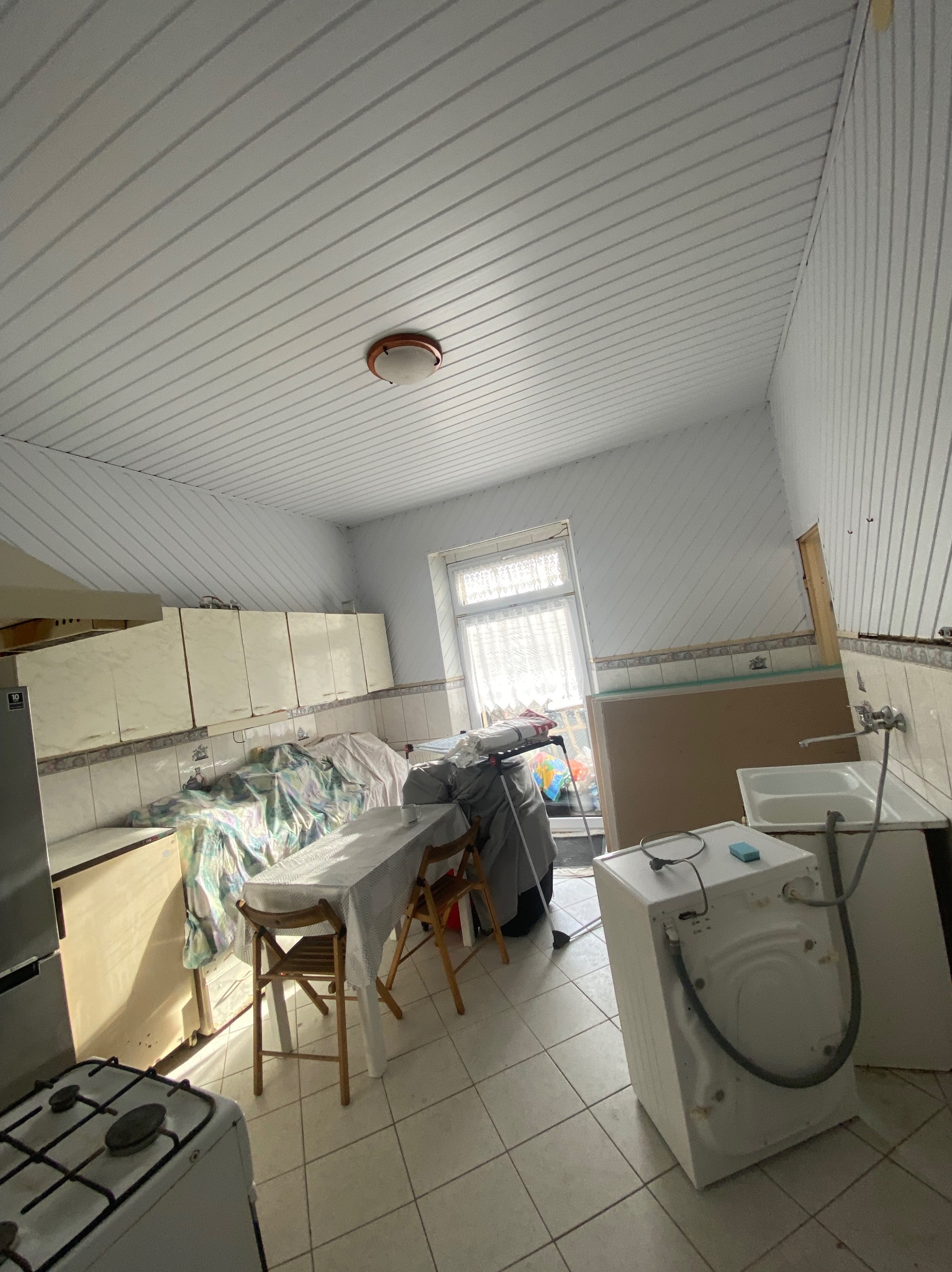 Kuchnia w trakcie prac, z zakrytymi meblami, widoczną pralką i elementami instalacji, sufit z paneli PCV, okno z firanką