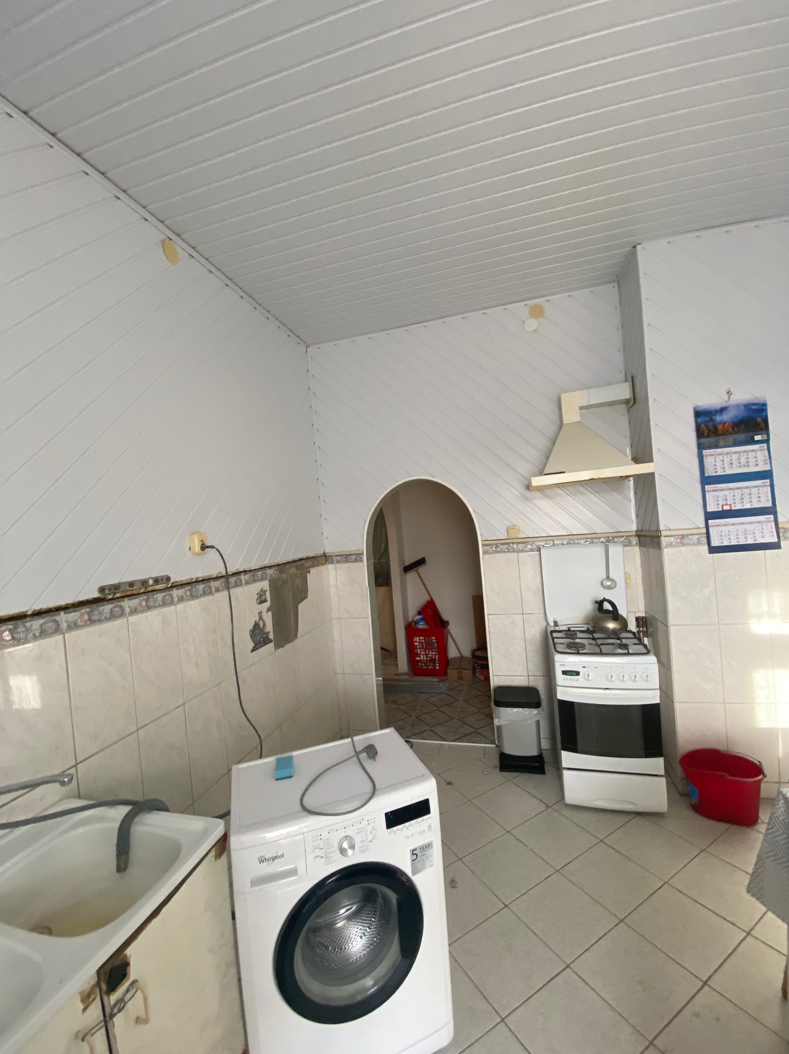 Kuchnia w trakcie remontu: widoczne panele ścienne i sufitowe, częściowo usunięte płytki, pralka, kuchenka i ślady po pracach wykończeniowych.