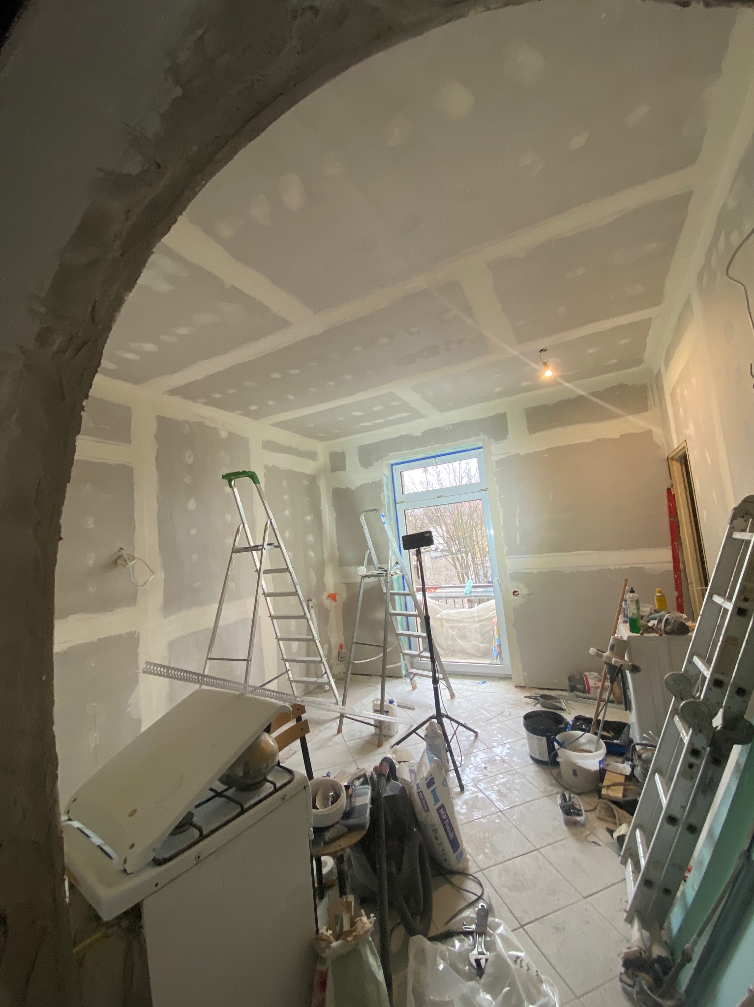 Wnętrze w trakcie szpachlowania i przygotowania do malowania, widoczne drabiny, narzędzia, worki z zaprawą, częściowo zakryty piec gazowy, okno zabezpieczone folią malarską.