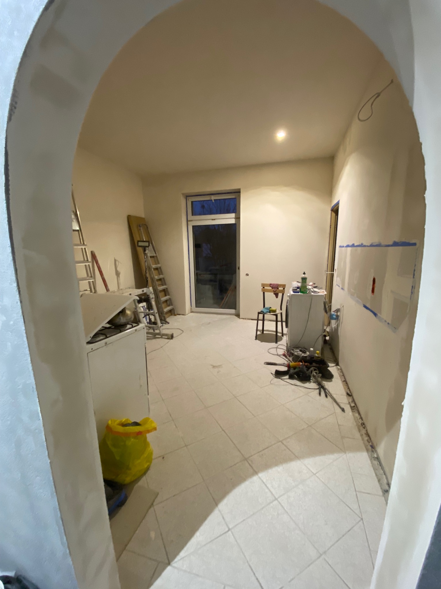 Wnętrze w trakcie prac wykończeniowych w Gliwicach: widoczne ściany przygotowane do malowania, drabiny, narzędzia oraz prowizoryczne wyposażenie, w tym pralka i krzesło. Uwagę zwraca żółta torba...