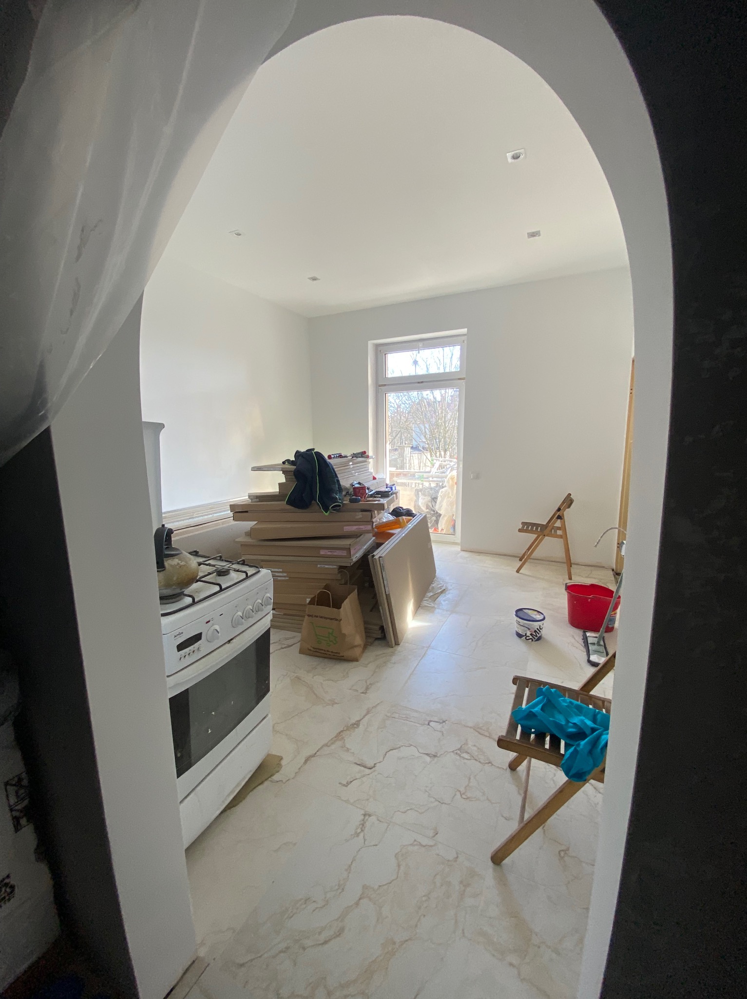 Widok przez łuk na remontowane wnętrze: białe ściany, marmurowe płytki, stos płyt kartonowo-gipsowych obok kuchenki, drabina, krzesła i akcesoria malarskie.