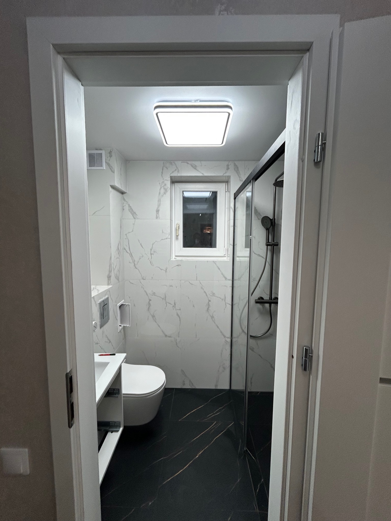 Wykończona łazienka w bieli i czerni z kabiną prysznicową, widoczna przez otwarte drzwi. Marmurowe płytki na ścianach, czarna podłoga z żyłkami, nowoczesne oświetlenie.