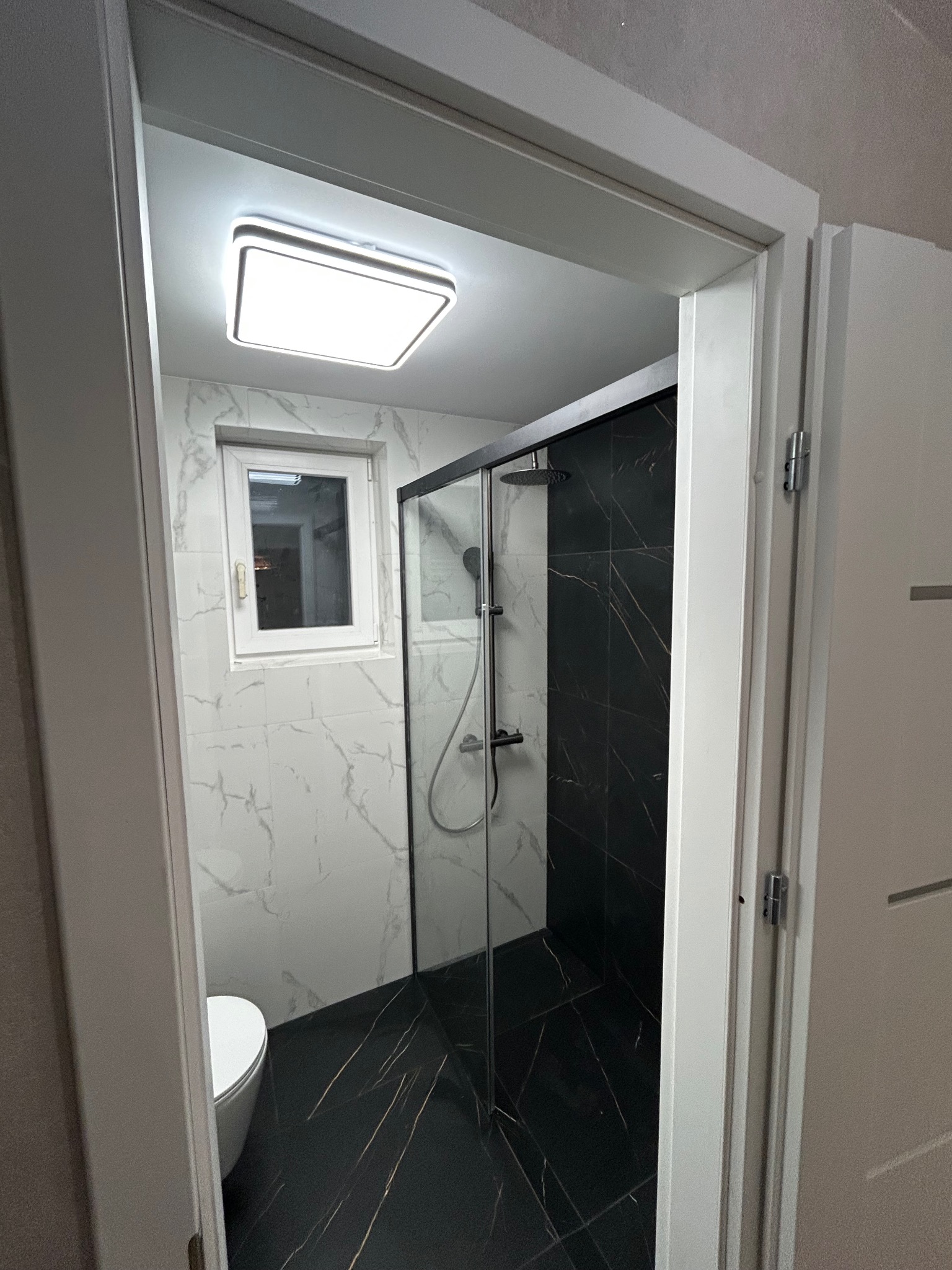 Nowoczesna łazienka po remoncie: kabina prysznicowa z czarnymi płytkami imitującymi marmur, białe marmurowe płytki na ścianach, oświetlenie sufitowe LED.