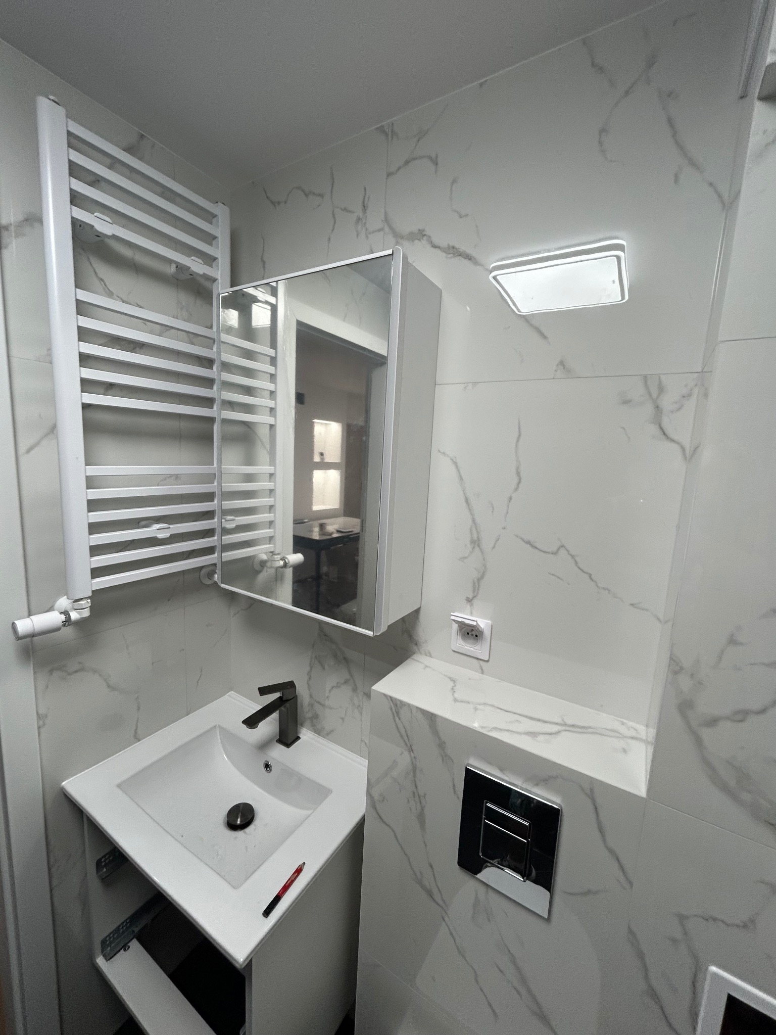 Wykończona łazienka w bieli i szarości z marmurkowym wzorem na płytkach. Widoczna umywalka z szafką, grzejnik drabinkowy, lustro z szafką i podświetlany sufit.