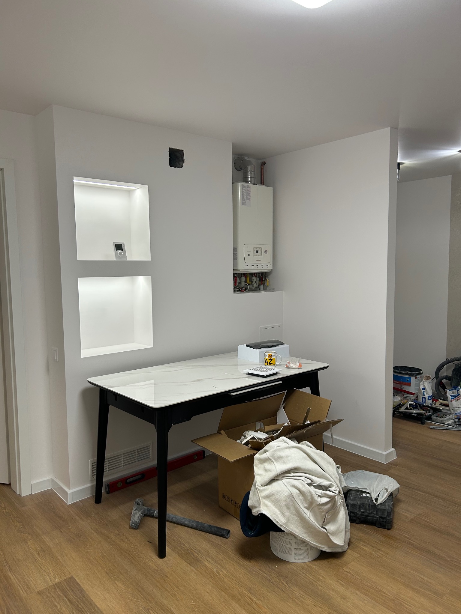 Białe ściany z wnękami podświetlonymi LED, wiszący piec gazowy, stół z narzędziami i akcesoriami malarskimi na podłodze. Wnętrze w trakcie wykańczania.
