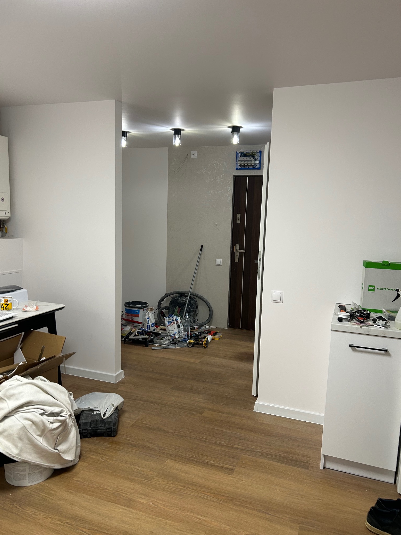 Wnętrze w trakcie prac wykończeniowych: widoczne narzędzia, puszki z farbą, rozłożone kable i zamontowana skrzynka elektryczna. Nowe oświetlenie sufitowe.