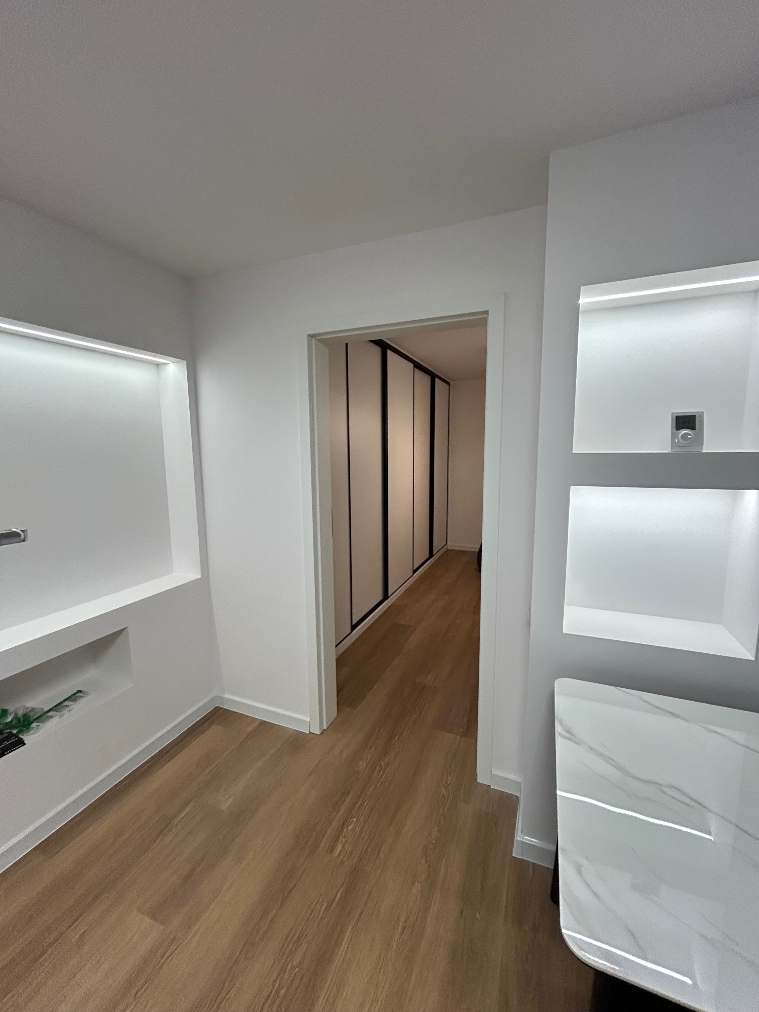 Białe wnętrze z drewnianą podłogą, wnękami z oświetleniem LED i szafą w zabudowie. Minimalistyczny design, jasne kolory, nowoczesne wykończenie.