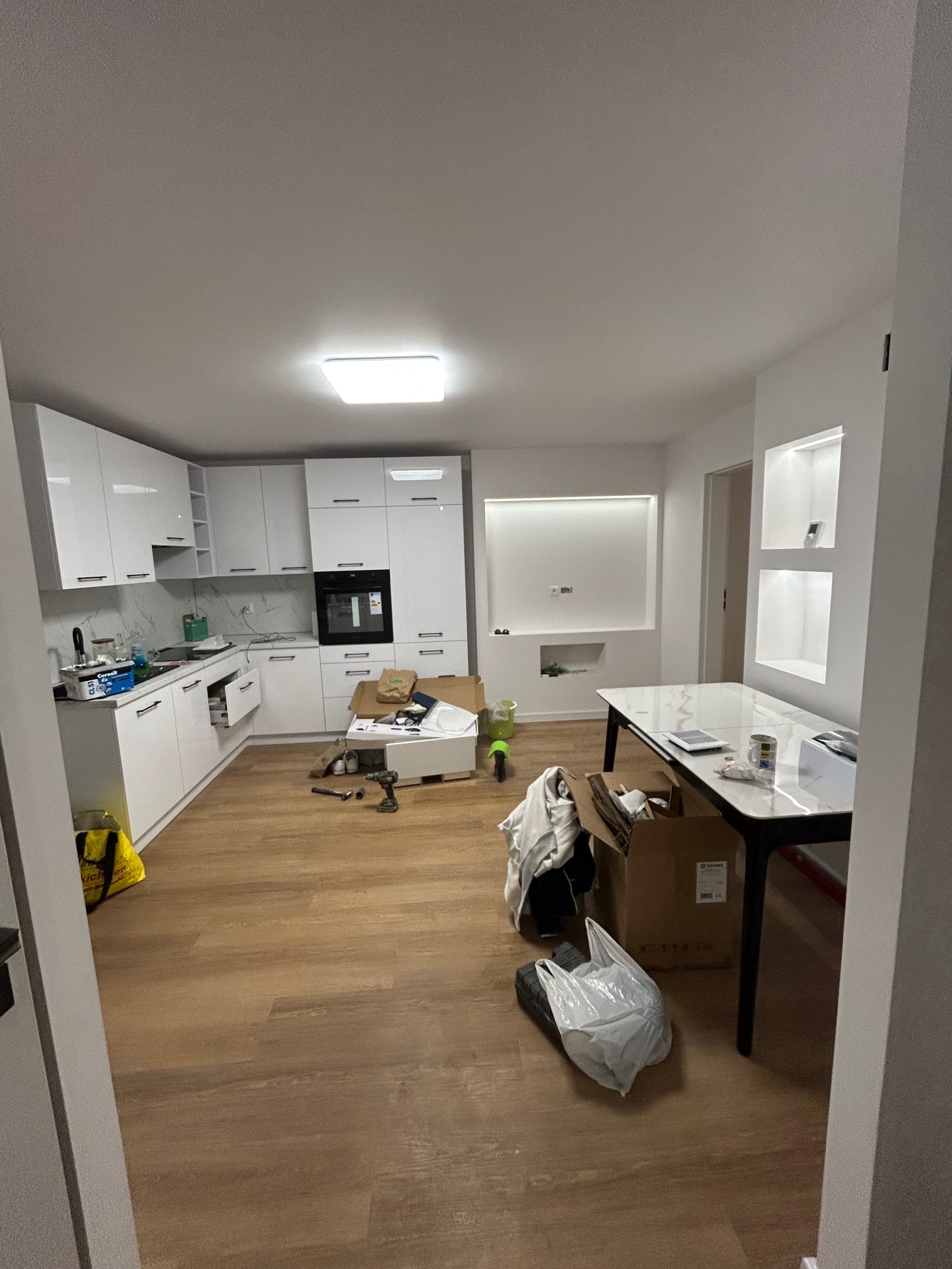 Jasne wnętrze kuchni w trakcie wykończenia: białe szafki, drewnopodobna podłoga, kartony i narzędzia sugerują trwające prace. Widoczne wnęki z oświetleniem.