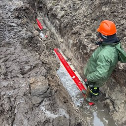 BEAVERBUD SPÓŁKA Z OGRANICZONĄ ODPOWIEDZIALNOŚCIĄ - Wykop z ułożonymi czerwonymi rurami karbowanymi, częściowo zalany wodą. Pracownik w kasku i zielonej kurtce nadzoruje ułożenie rur w błotnistym terenie.