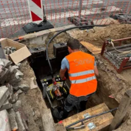 Pracownik w wykopie, w pomarańczowej kamizelce, obsługuje maszynę. Widoczne zabezpieczenia wykopu, kostka brukowa i elementy instalacji.
