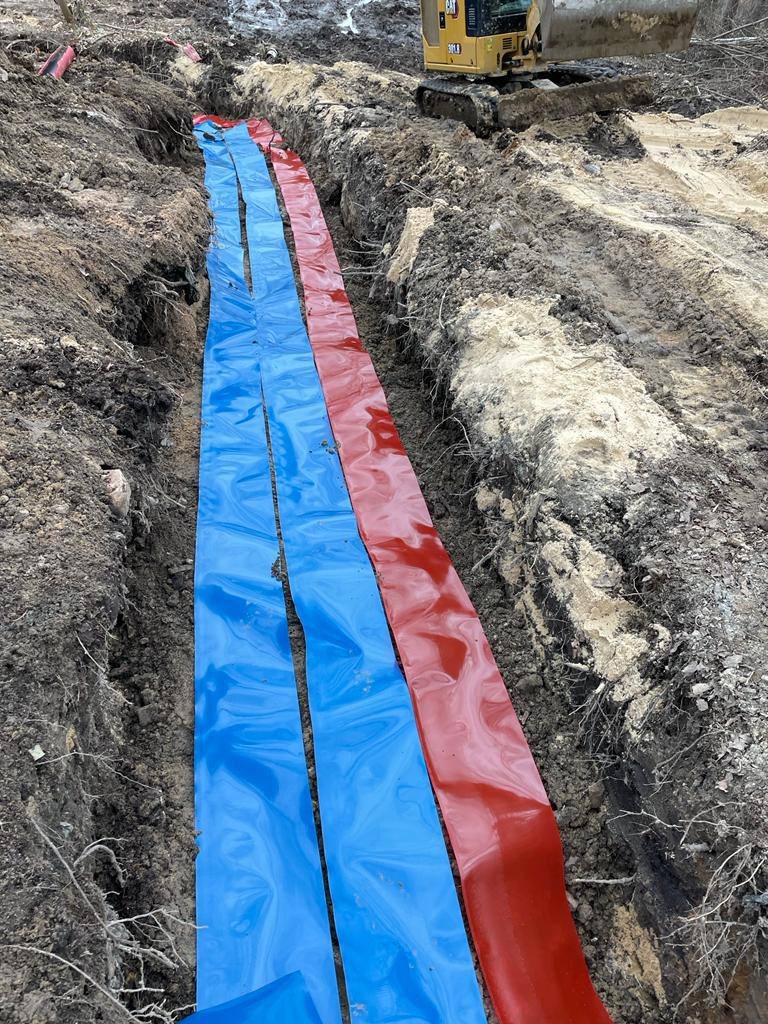 Wykopany rów z ułożonymi niebieskimi i czerwonymi taśmami zabezpieczającymi, w tle koparka CAT na nierównym terenie.