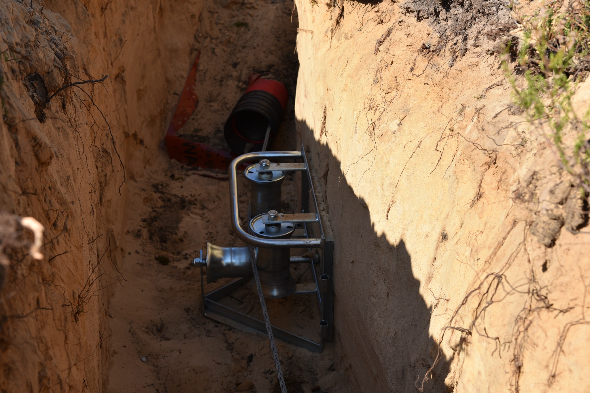 Wąski wykop w piaszczystej glebie z widocznym metalowym oprzyrządowaniem do przeciągania kabli i fragmentem rury karbowanej w tle.