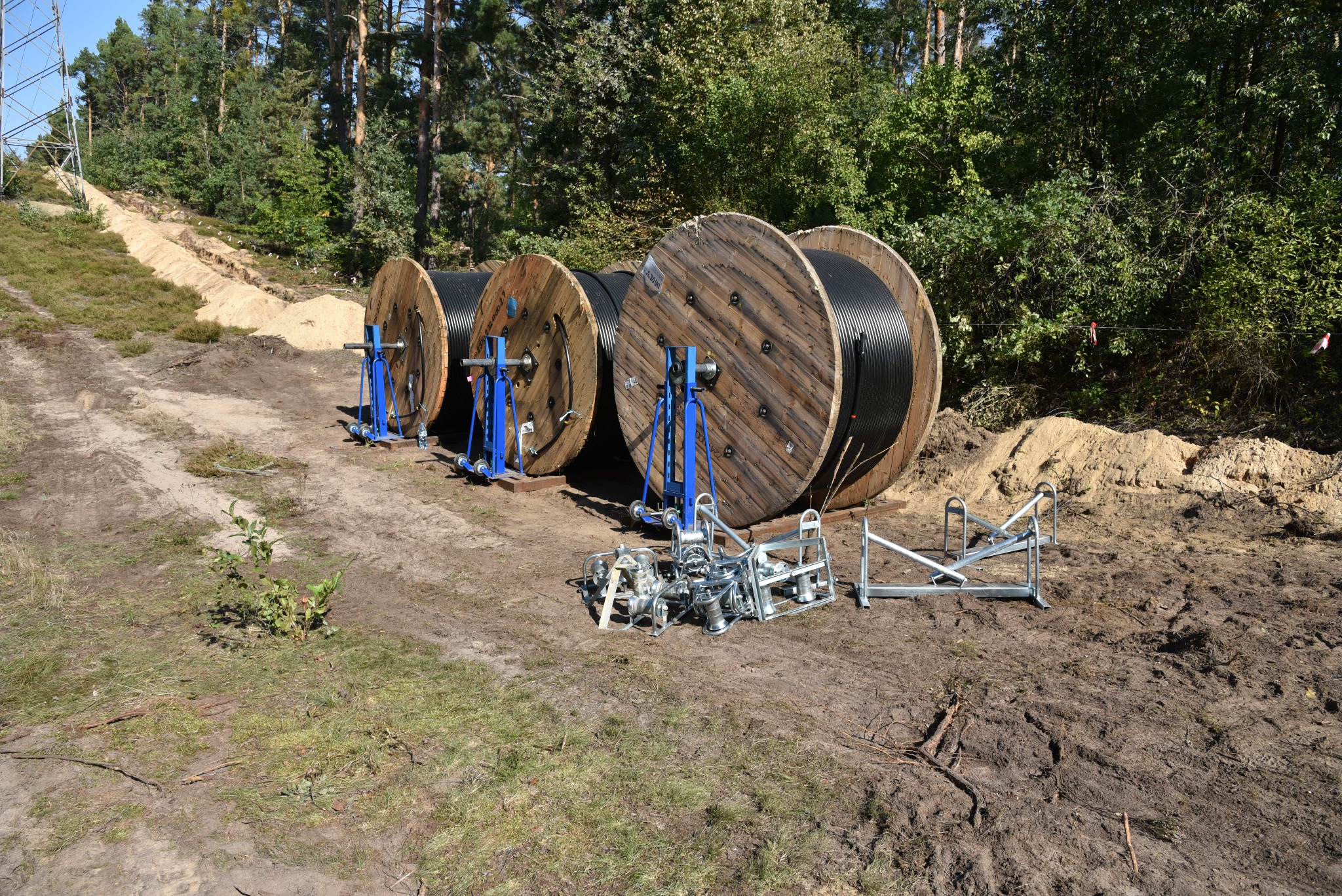 Trzy drewniane szpule z czarnym kablem na nieutwardzonym terenie, podtrzymywane przez niebieskie stojaki; wykop ziemny widoczny w tle, sugerujący prace instalacyjne.